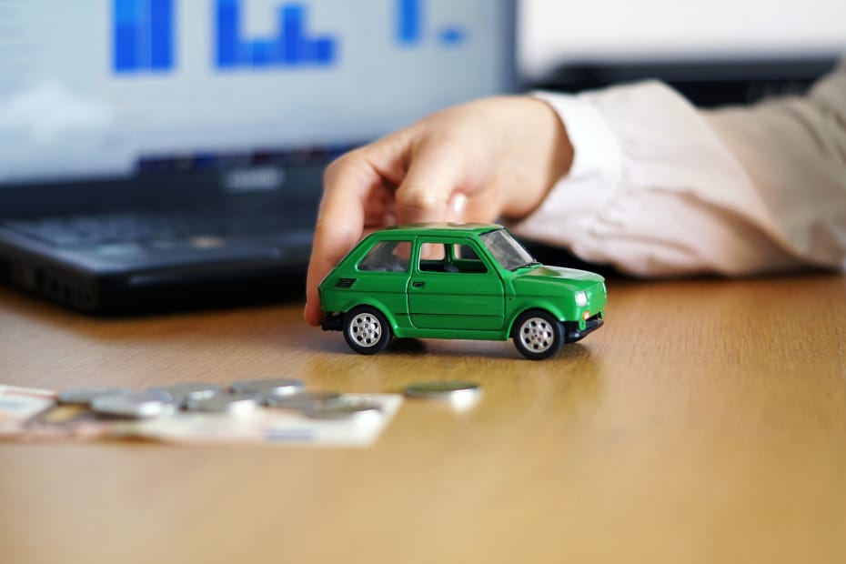 Hand holding a small green toy car over a desk with coins and a laptop, symbolizing getting the best scrap car prices online.